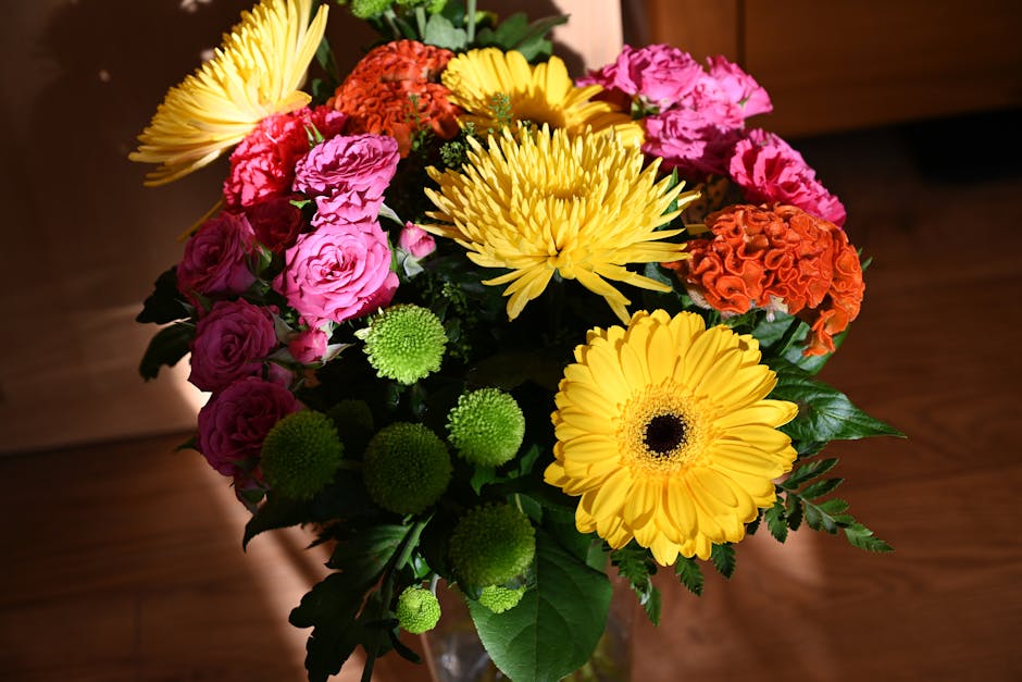 A vibrant floral arrangement featuring large, bright yellow gerbera daisies with dark centers, pink roses with soft, layered petals, orange carnations with ruffled blooms, and green button chrysanthem