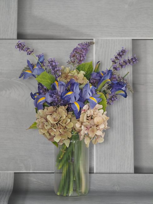 A tall, clear glass vase filled with a vibrant floral arrangement featuring large, pale pink hydrangeas, deep blue irises with yellow accents, and sprigs of purple lavender. The bouquet is arranged wi