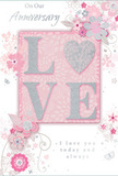 The image shows a softly romantic anniversary card, beautifully styled in pastel pinks and shimmering neutrals that would pair perfectly with a vase of fresh flowers in a Watford living room. At the heart of the design, the word LOVE stands out in prominent, raised letters, covered with fine glitter that blends blush-pink, pearlescent white, and delicate hints of silver. The texture is clearly visible, suggesting a pleasant, slightly rough feel when touched, much like brushing your fingers over the centre of a fresh chrysanthemum. Encircling this central word is a loose wreath of stylised flowers, curling vines, and slender leaves drifting across a pale pink background. Each blossom is depicted with fine, graceful lines in grey and silver, then shaded with the softest tones of pink and mauve, so the flowers appear almost like a watercolour sketch-a florist's first, inspired draft of an anniversary bouquet. Subtle glimmers of glitter accent selected petals and swirling lines, adding a sense of festivity similar to the sparkle of glassware on a table set for an anniversary meal near Watford's town centre. The overall composition remains light and unhurried, leaving breathable space around the artwork to keep the mood calm and intimate. A flowing, hand-lettered-style script delivers a heartfelt message about shared love and commitment, the words today and always nestling into the design with natural ease. The script's soft grey ink harmonises with the floral outlines, avoiding harsh contrast and keeping the card's atmosphere gentle and affectionate. Altogether, this card feels like a tender, tactile companion to a bouquet-an elegant way to mark another year together, whether it's delivered with flowers to a flat near the high street or a quiet home on the outskirts of town.